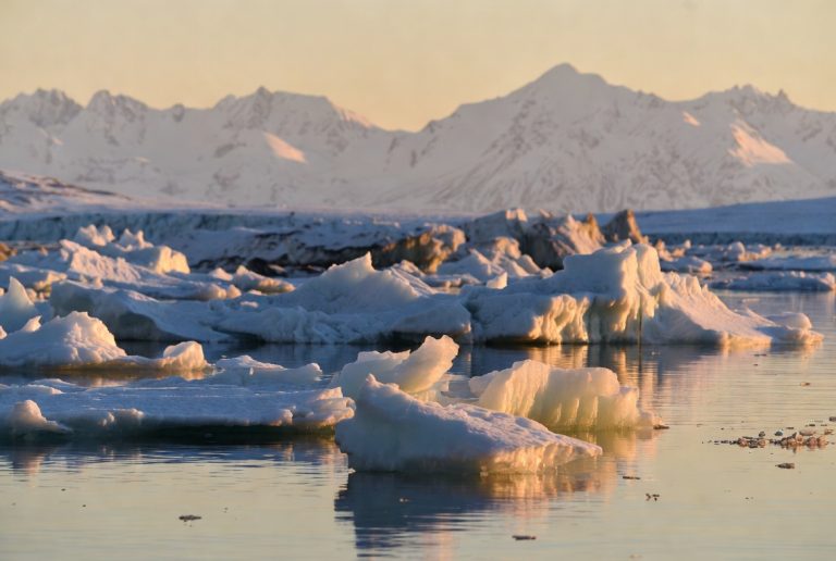 Realistic Arctic sea ice floes floating in calm water at golden hour with snow-covered mountains in the background.