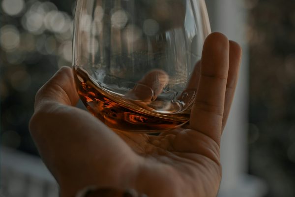 A man’s hand holding a glass of whiskey on an outdoor balcony.
