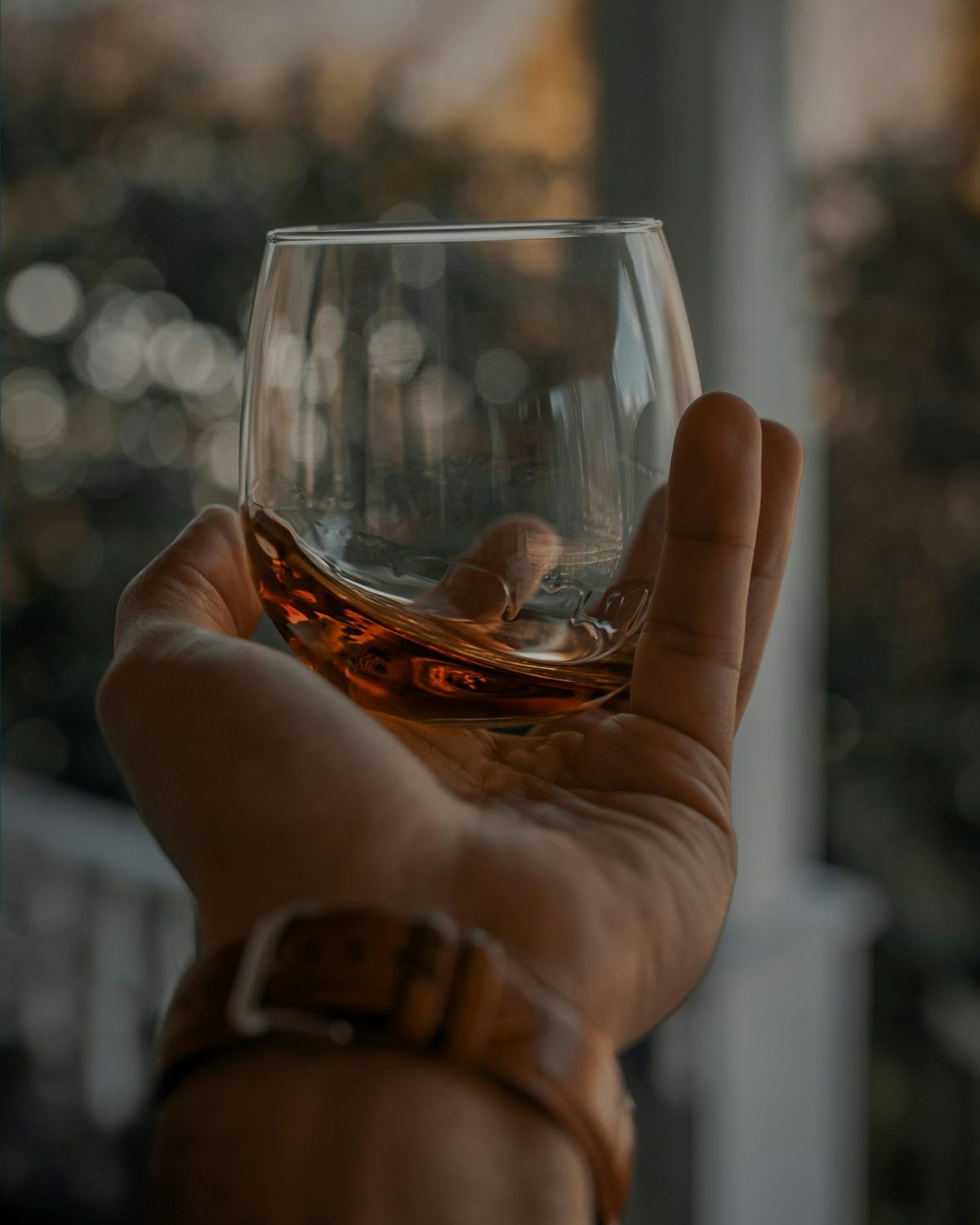 A man’s hand holding a glass of whiskey on an outdoor balcony.
