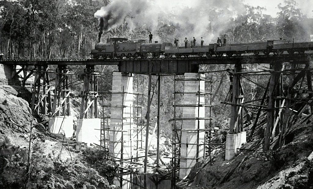 black and white Image of a steam train on top of a railway bridge.