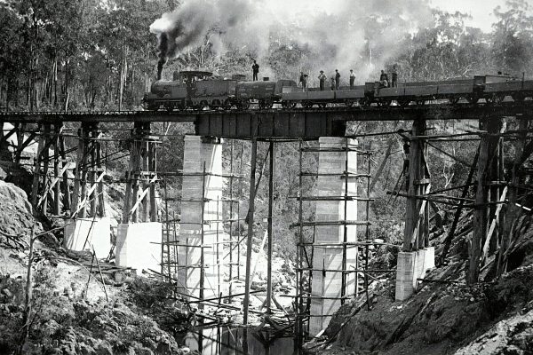 black and white Image of a steam train on top of a railway bridge.