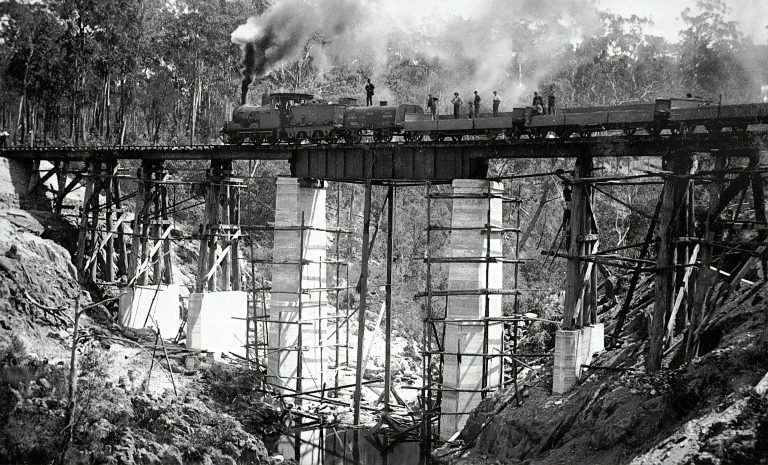 black and white Image of a steam train on top of a railway bridge.