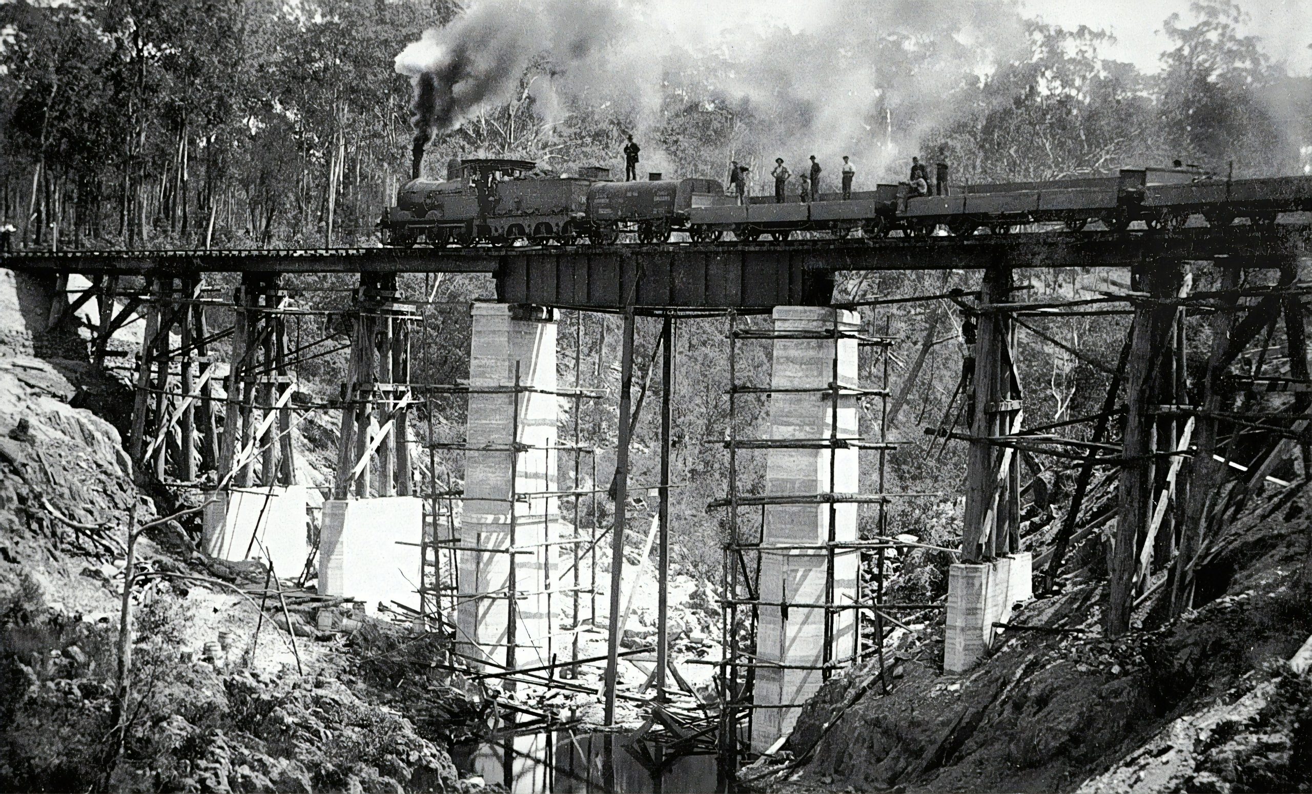 black and white Image of a steam train on top of a railway bridge.