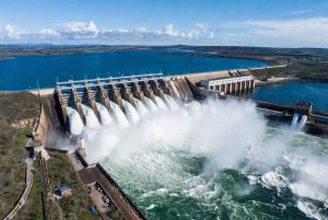 Aerial view of a massive hydroelectric dam releasing powerful torrents of water through its spillways, creating dramatic white foam and spray in the river below. A large blue reservoir stretches behind the concrete structure under a clear sunny sky