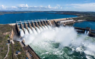 Aerial view of a massive hydroelectric dam releasing powerful torrents of water through its spillways, creating dramatic white foam and spray in the river below. A large blue reservoir stretches behind the concrete structure under a clear sunny sky