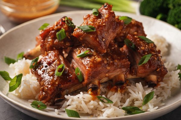 Tender baby back pork ribs coated in a glossy, sticky honey garlic glaze, piled over fluffy white jasmine rice and garnished with sliced green onions and sesame seeds. A small bowl of extra glaze and steamed broccoli are visible in the soft background.