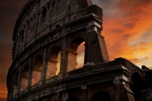 Dramatic sunset view of the ancient Colosseum in Rome, its massive stone arches and towering facade glowing against a fiery orange and red sky, capturing the epic grandeur of the Roman Empire.