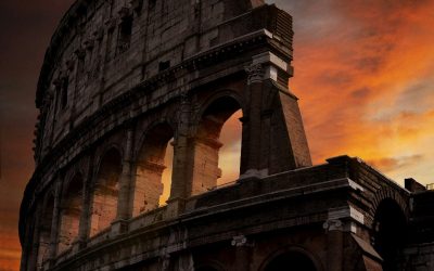 Dramatic sunset view of the ancient Colosseum in Rome, its massive stone arches and towering facade glowing against a fiery orange and red sky, capturing the epic grandeur of the Roman Empire.