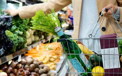 Shopper carrying a wire shopping basket with a green glass bottle, fresh celery, lemon, zucchini, and bread while reaching for vegetables in the supermarket produce section.