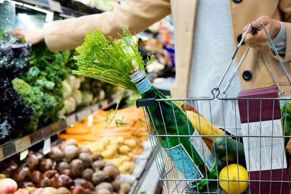 Shopper carrying a wire shopping basket with a green glass bottle, fresh celery, lemon, zucchini, and bread while reaching for vegetables in the supermarket produce section.