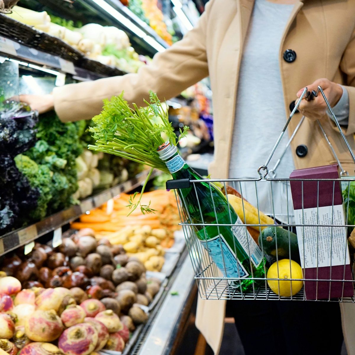 Shopper carrying a wire shopping basket with a green glass bottle, fresh celery, lemon, zucchini, and bread while reaching for vegetables in the supermarket produce section.