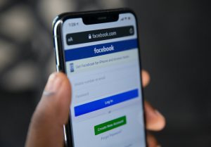 Close-up of a hand holding a smartphone displaying the Facebook login page, with fields for mobile number or email and password, a blue Log In button, and a green Create New Account button.