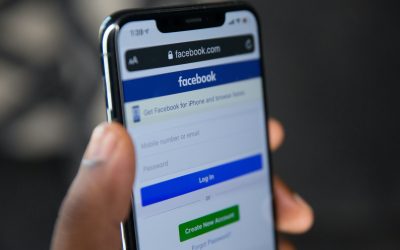 Close-up of a hand holding a smartphone displaying the Facebook login page, with fields for mobile number or email and password, a blue Log In button, and a green Create New Account button.