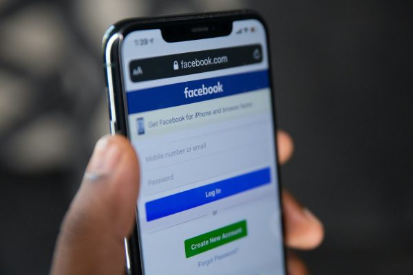 Close-up of a hand holding a smartphone displaying the Facebook login page, with fields for mobile number or email and password, a blue Log In button, and a green Create New Account button.