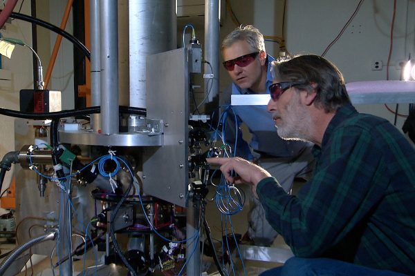 Two scientists standing beside the tall NIST-F2 cesium fountain atomic clock in a laboratory.