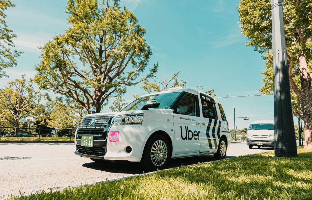 White Uber taxi parked on a sunny street beside grass and trees, with another vehicle in the background.