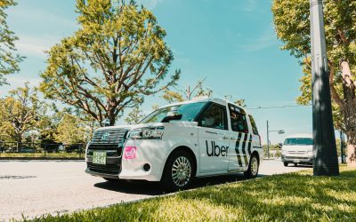 White Uber taxi parked on a sunny street beside grass and trees, with another vehicle in the background.