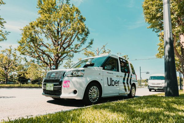 White Uber taxi parked on a sunny street beside grass and trees, with another vehicle in the background.