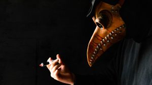 Close-up of a leather bird-beak plague-doctor mask as a gloved hand holds a syringe against a black background.