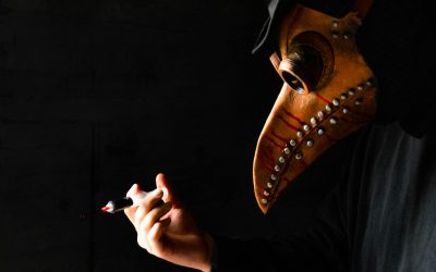 Close-up of a leather bird-beak plague-doctor mask as a gloved hand holds a syringe against a black background.