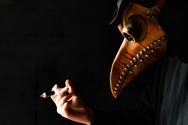 Close-up of a leather bird-beak plague-doctor mask as a gloved hand holds a syringe against a black background.