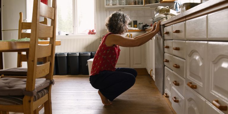 A woman with curly gray hair crouches barefoot in a sunlit kitchen and reaches toward a built-in dishwasher under the counter, with wooden chairs and a table in the foreground.