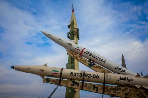 Iranian missiles on display outdoors, including one upright launcher and two missiles on camouflage rails marked with Iranian flags and serial numbers.