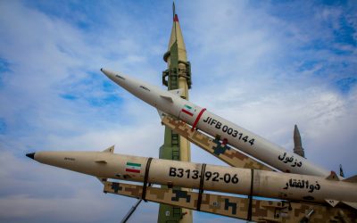Iranian missiles on display outdoors, including one upright launcher and two missiles on camouflage rails marked with Iranian flags and serial numbers.