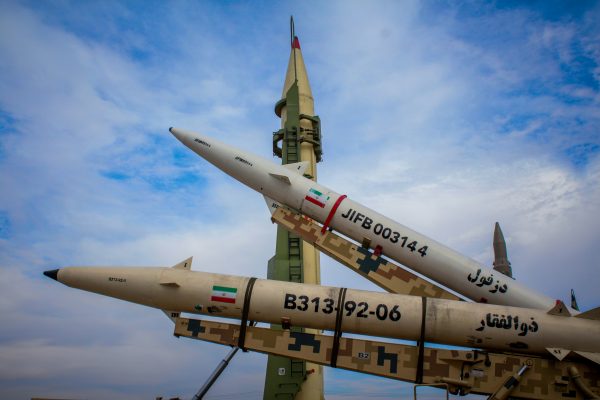 Iranian missiles on display outdoors, including one upright launcher and two missiles on camouflage rails marked with Iranian flags and serial numbers.
