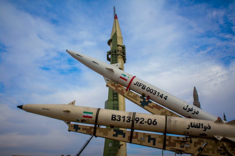 Iranian missiles on display outdoors, including one upright launcher and two missiles on camouflage rails marked with Iranian flags and serial numbers.