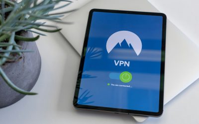 Overhead view of a tablet resting on a closed silver laptop, with a blue VPN screen showing a mountain logo, an on toggle, and the status “You are connected”.
