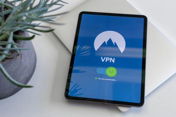Overhead view of a tablet resting on a closed silver laptop, with a blue VPN screen showing a mountain logo, an on toggle, and the status “You are connected”.