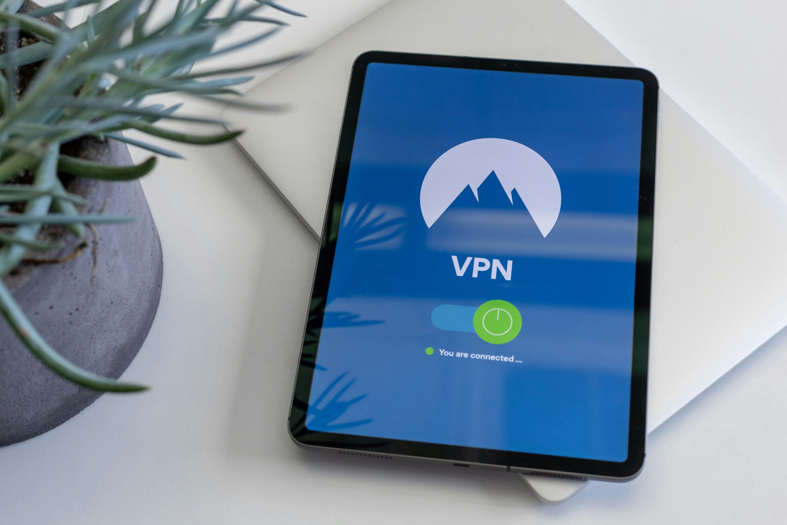 Overhead view of a tablet resting on a closed silver laptop, with a blue VPN screen showing a mountain logo, an on toggle, and the status “You are connected”.