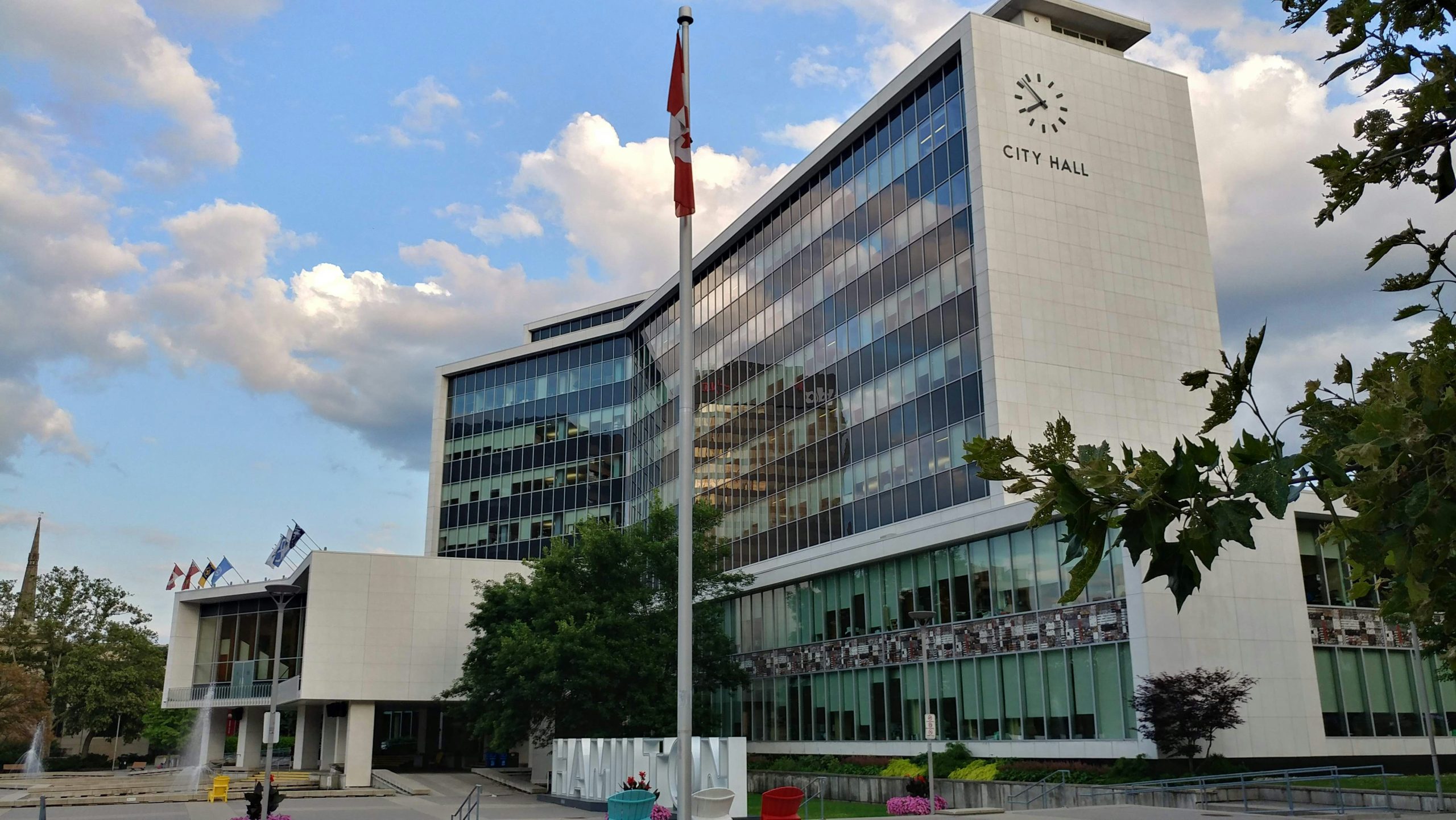 Hamilton City Hall, a white office tower marked “CITY HALL,” with a Canadian flag in front.