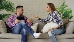 Couple on beige sofa in a tense conversation; plants and white brick wall behind them.