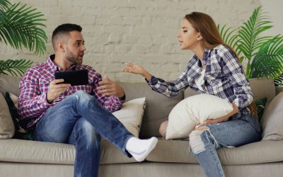 Couple on beige sofa in a tense conversation; plants and white brick wall behind them.