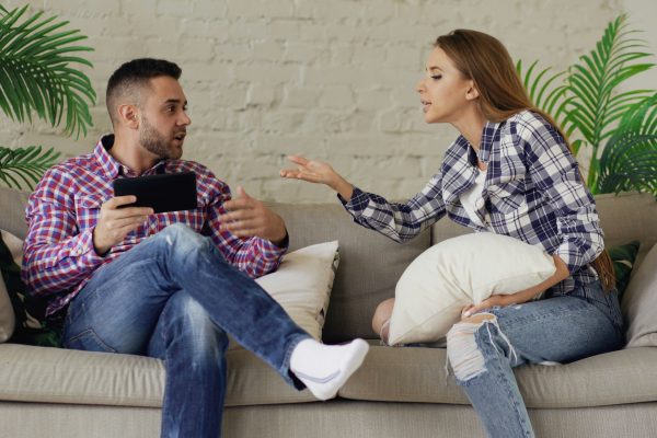 Couple on beige sofa in a tense conversation; plants and white brick wall behind them.