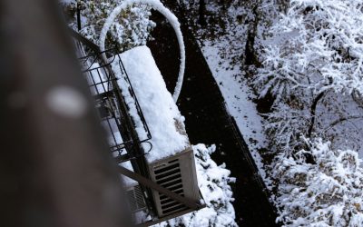 Snow-covered mini-split outdoor unit mounted on a balcony, with iced refrigerant lines and snow-laden trees below.