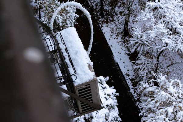 Snow-covered mini-split outdoor unit mounted on a balcony, with iced refrigerant lines and snow-laden trees below.