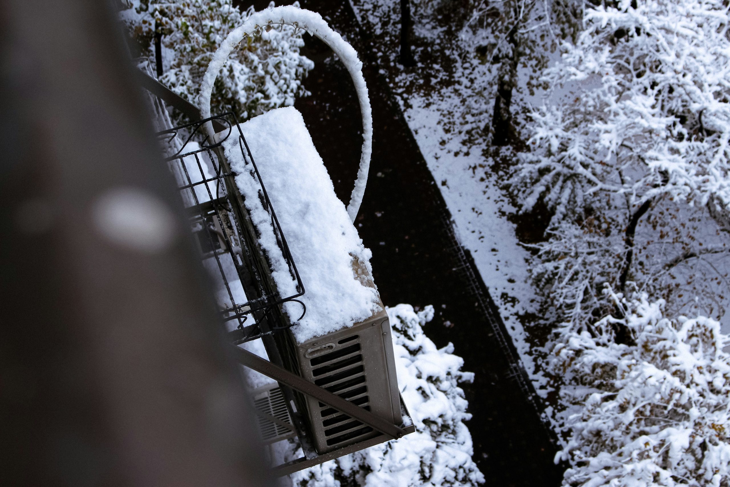 Snow-covered mini-split outdoor unit mounted on a balcony, with iced refrigerant lines and snow-laden trees below.